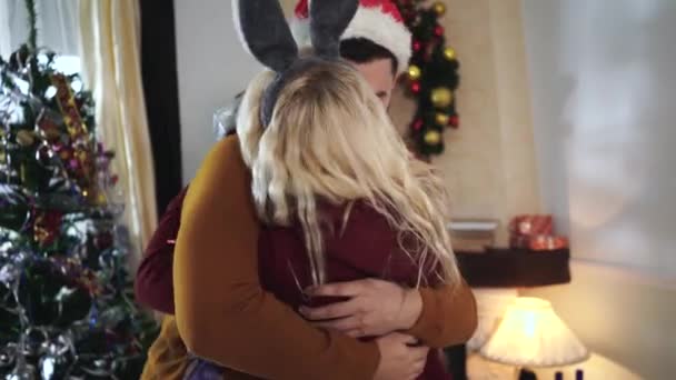 Close-up of Caucasian man and woman hugging in front of Christmas tree. Blond wife holding gift box and smiling. Happy couple spending New Years eve together at home. Holidays season, celebration. - Filmati, video