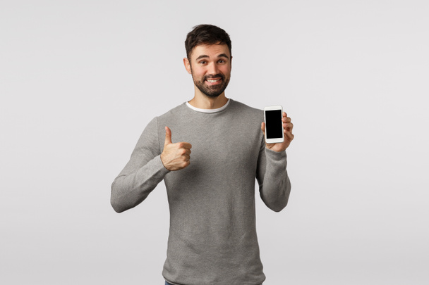 Friendly handsome and delighted bearded man in grey sweater recommend use application or like new messanger, shopping site, holding smartphone, show thumb-up in approval, like, smiling pleased - Photo, Image