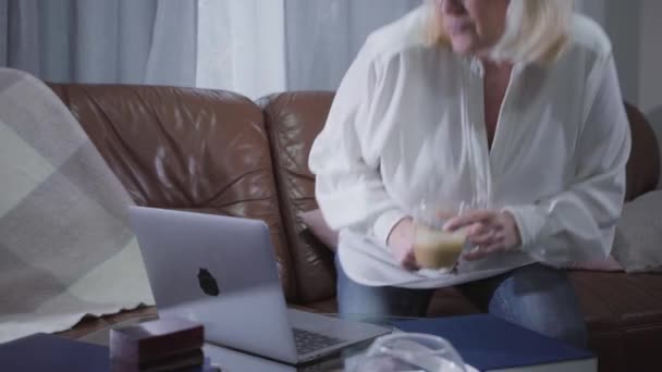 Positive Caucasian retiree sitting on sofa with coffee cup and looking at laptop screen. Senior blond woman surfing internet at home. Pastime, leisure, social networks. - Metraje, vídeo