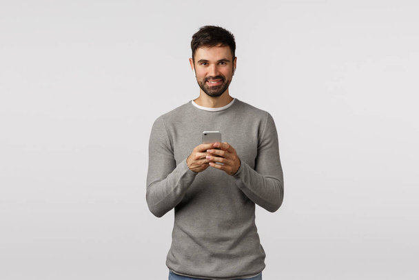 Attractive friendly-looking caucasian man in grey sweater, holding smartphone, wear wireless headphones, using earphones to listen music, watch video in public place, stand white background - Photo, Image