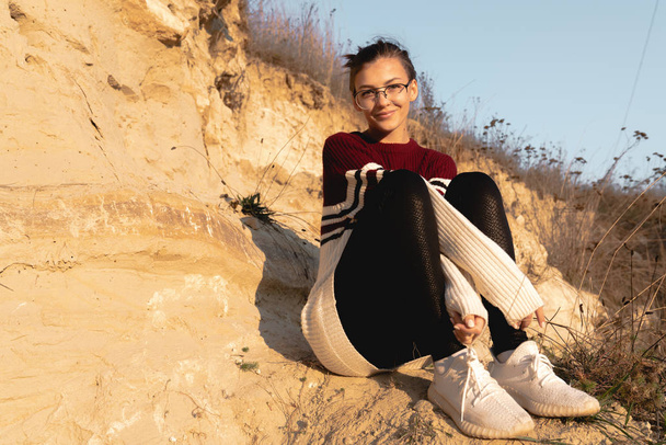 Gyönyörű barna lány szemüvegben, piros pulóverben, leül a földre a homokbánya mellett, és a kamerába néz. - Fotó, kép
