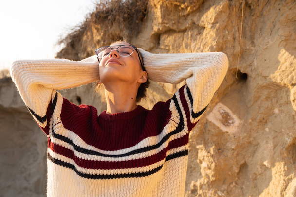 Close up retrato da bela menina morena em óculos e suéter fica ao sol com os olhos fechados e mãos levantadas
 - Foto, Imagem