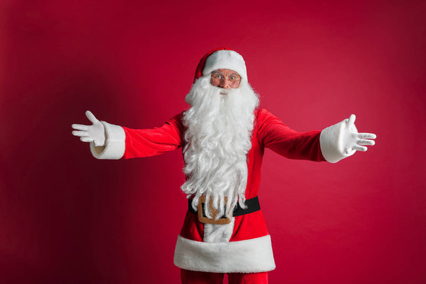 Middle age handsome man wearing Santa costume standing over isolated red background smiling cheerful with open arms as friendly welcome, positive and confident greetings - Foto, afbeelding