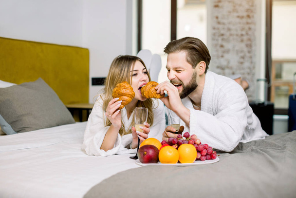Lovers celebrating an engagement or honeymoon with fresh fruits, croissants and champagne in bed, hotel room. Man feeding woman with croissant - Fotografie, Obrázek