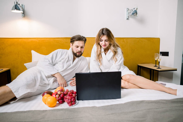 Relaxed couple working on laptop in bed. Couple of freelancers, lovers, business people. Couple wearing hotel bathrobes drinking champagne and eating fruits in bed and working - Zdjęcie, obraz