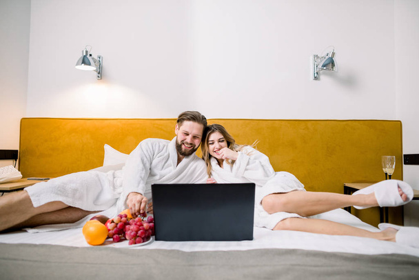 Front view of sweet married couple lying in bed, looking at laptop and smiling. Husband and wife in love wearing white bathrobes in modern hotel room - Φωτογραφία, εικόνα