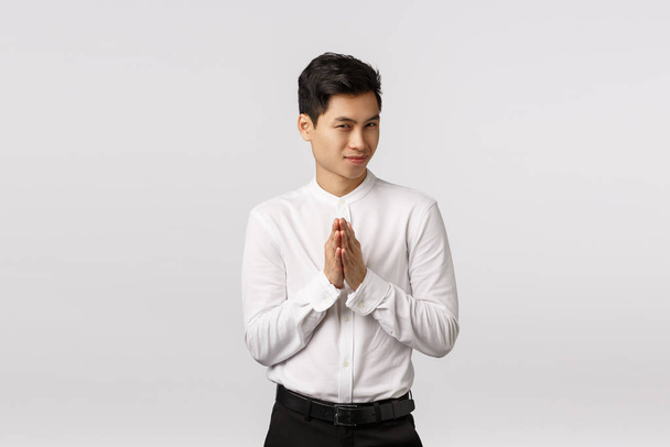 Mysterious and devious young good-looking asian businessman in white shirt, pants, rub hands and squinting sly, have secret evil plan, relish good deal, make money, standing white background - Photo, Image