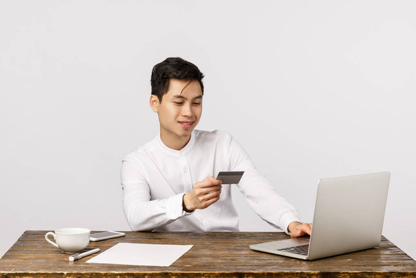 起業家、金融、採用の概念。ハンサムなアジアのビジネスマン25秒、テーブルに座って、クレジットカードの笑顔を保持し、オンライン注文を行い、ノートパソコンに銀行番号を挿入し、コーヒーを飲む - 写真・画像