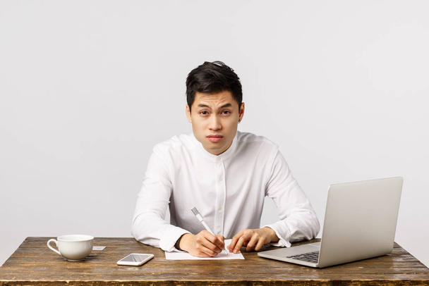 Tired and skeptical young asian male student trying take notes and catch lector speech, looking exhausted with sleepy eyes, squinting to clear view, sit desk with laptop, drink coffee, hardworking - Zdjęcie, obraz