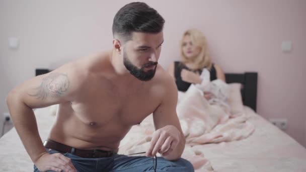 Close-up portrait of young bearded Caucasian man with tattoo on shoulder sitting on bed, taking off eyeglasses, and shaking head. Upset blond woman lying at the background. Intimate problems. - Filmati, video