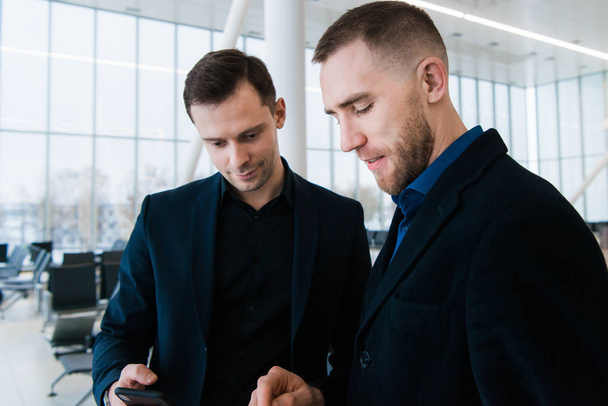 Two handsome businessmen in airport, discussing important project and looking at phone screen. - Foto, imagen