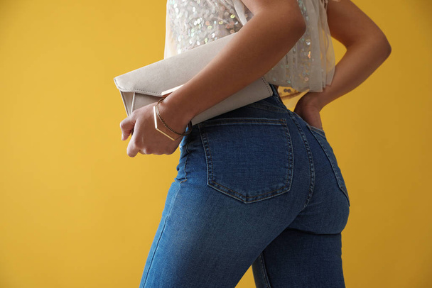 Woman in jeans with clutch purse on yellow background, closeup - Zdjęcie, obraz