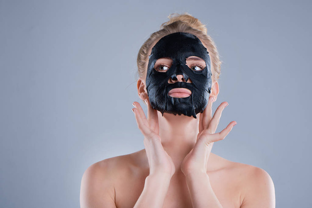 menina com ombros nus posando em fundo cinza com máscara facial preta, conceito de cuidados com a pele, foto de beleza, tratamento de pele, máscara de pele hidratante, sorrindo
. - Foto, Imagem