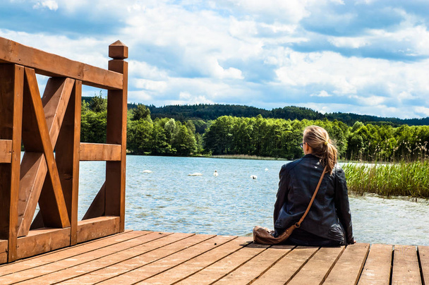 Krajobraz drewnianego molo nad pięknym jeziorem w lecie. Tylny widok kobiety siedzącej na pomoście otoczonej naturą. Urlop i urlop. - Zdjęcie, obraz