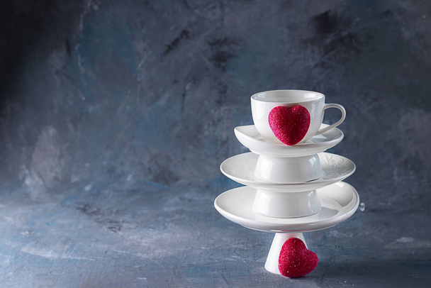 White saucer pyramid with cup of tea on top and two red hearts candy on it Dark background. Creative concept, love, relations, gift, celebrate, restaurant, food, St. Valentine's day. Square copy space - Photo, image
