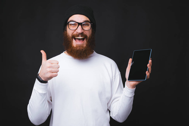 Veselý mladý vousatý muž drží tabletu a ukazuje palce nahoru - Fotografie, Obrázek
