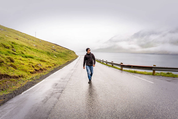 Young traveller walking y empty road after rain, green mountains, fjord and overcast sky on horizon. Faroe Islands, Denmark. - Photo, Image