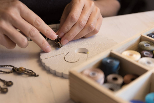 Lifestyle concept, werken vanuit huis om je leven opnieuw uit te vinden: close-up van de vrouw handen maken macrame geknoopte sieraden met stenen kralen en gereedschappen op lichte houten tafel - Foto, afbeelding