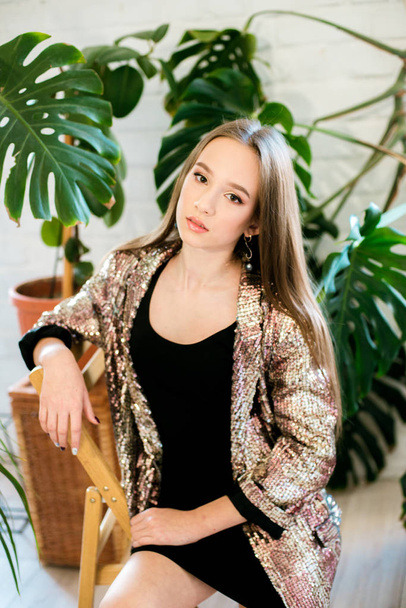 Teenager girl with long hair in a black dress and a sparkling jacket among tropical plants in tudia. Evening fashion - Foto, Imagen