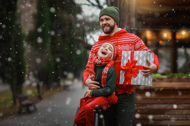 Dad with his son in bright holiday clothes posing in front of the camera on the street and holding a box with a gift in their hands. - Фото, изображение