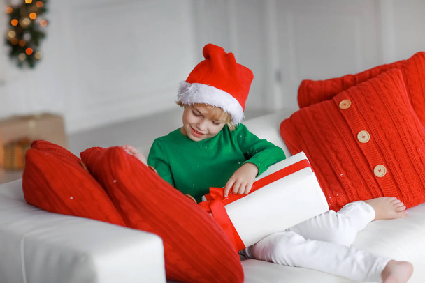 A boy in a santaclaus cap and a green sweater sits on a sofa and holds a white box with a gift - Фото, зображення