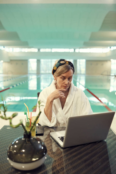 Portrait taille haute d'une femme âgée riche utilisant un ordinateur portable tout en se relaxant dans un spa de luxe avec piscine en arrière-plan
 - Photo, image
