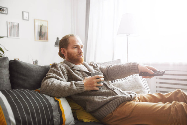 Porträt eines bärtigen Mannes mittleren Alters, der zu Hause fernsieht und die Kanäle per Fernbedienung schaltet, während er es sich auf der bequemen Couch gemütlich macht - Foto, Bild