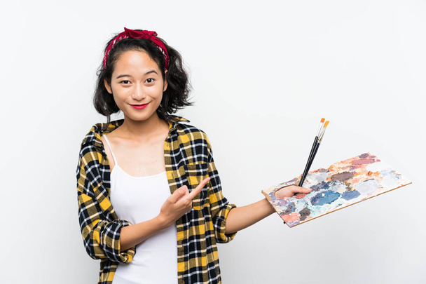 Joven artista mujer sosteniendo una paleta sobre fondo blanco aislado y señalándolo
 - Foto, Imagen