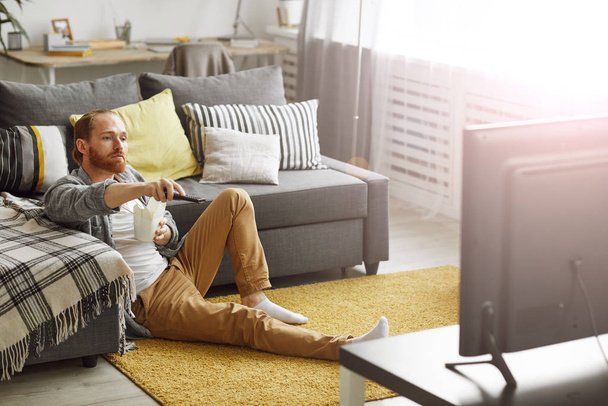 Full length πορτρέτο του γενειοφόρου ανθρώπου αλλαγή καναλιών, ενώ βλέποντας τηλεόραση στο σπίτι σε bachelors pad, αντίγραφο χώρου - Φωτογραφία, εικόνα