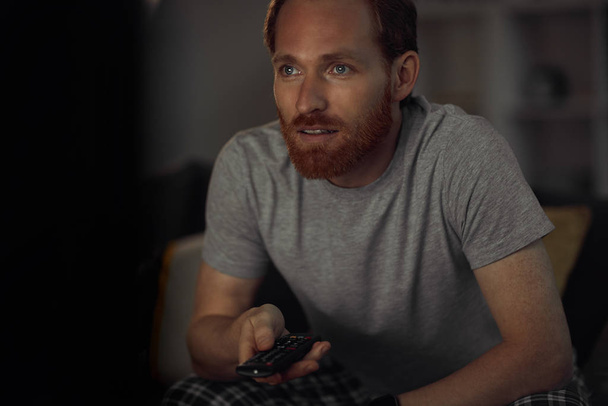 Porträt eines bärtigen erwachsenen Mannes, der nachts fernsieht, während er im dunklen Raum auf der Couch sitzt und die Kanäle wechselt, Kopierraum - Foto, Bild