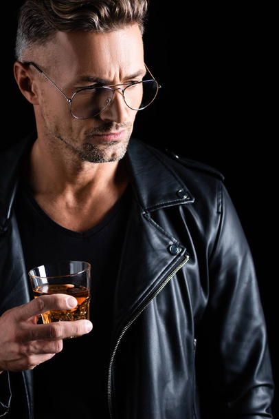 Handsome man in leather jacket holding glass of whiskey isolated on black - Фото, изображение
