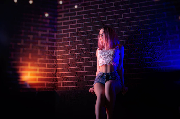 Μια λεπτή κοπέλα στον τοίχο με κοντά σορτς και πίσω δηλητηριώδες φως. - Φωτογραφία, εικόνα