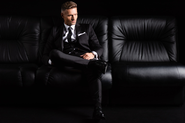 Confident businessman in formal wear sitting on couch isolated on black - Φωτογραφία, εικόνα