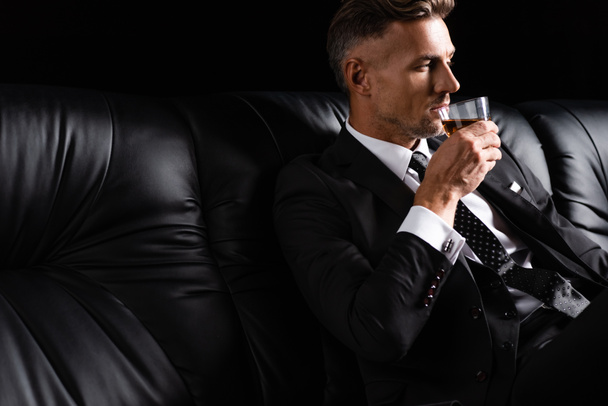 Handsome businessman drinking whiskey while sitting on couch isolated on black - Fotografie, Obrázek