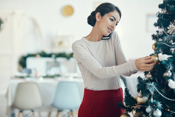 Porträt einer eleganten jungen Frau, die telefoniert, während sie zu Hause den Weihnachtsbaum schmückt - Foto, Bild
