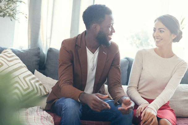 Portrait of mixed-race couple smiling at each other while talking sitting on cozy sofa at home, copy space - Foto, immagini