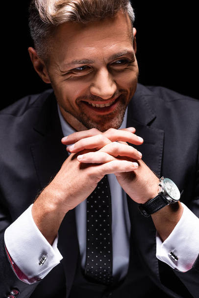 Glimlachende zakenman kijkt weg terwijl gekruiste vingers geïsoleerd op zwart  - Foto, afbeelding