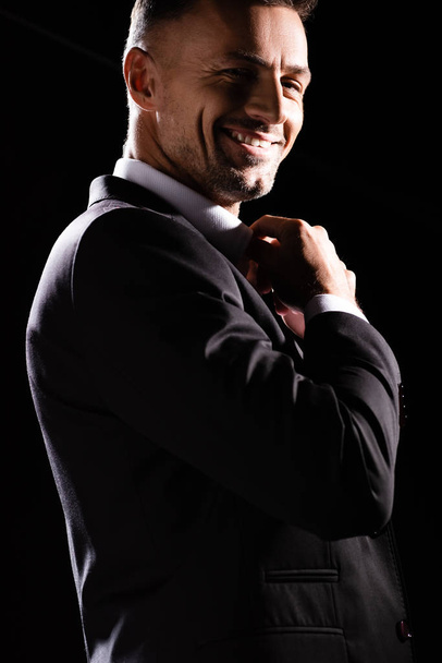 Side view of smiling businessman adjusting collar on shirt and looking at camera isolated on black  - Foto, Bild