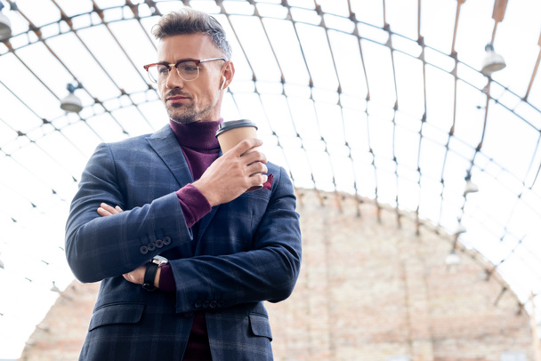 Low angle view of businessman in wireless earphones holding coffee to go on urban street - Photo, Image