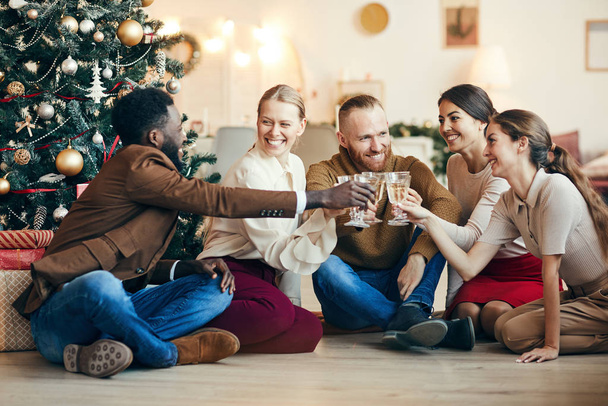 Plný portrét přátel cinkajících sklenicemi šampaňského sedících u vánočního stromku během párty, kopírovací prostor - Fotografie, Obrázek