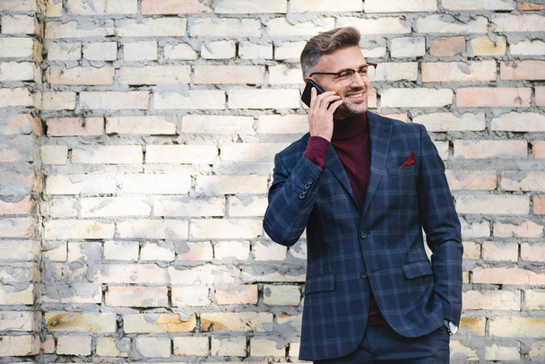 Улыбающийся бизнесмен разговаривает на смартфоне с кирпичной стеной на заднем плане
 - Фото, изображение