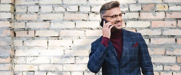 Panoramic shot of smiling businessman talking on smartphone with brick wall at background - Фото, зображення