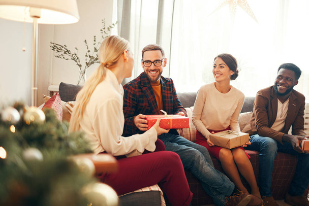 Grupo de adultos elegantes que intercambian regalos en Navidad, se centran en el hombre barbudo sonriendo felizmente sosteniendo la caja de regalo roja, espacio para copiar
 - Foto, Imagen