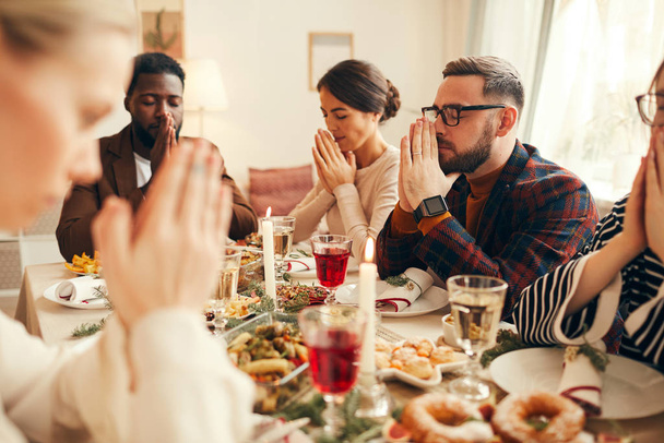 Grupo multiétnico de adultos elegantes que rezan sentados en la mesa de la cena durante el banquete de Navidad
 - Foto, Imagen