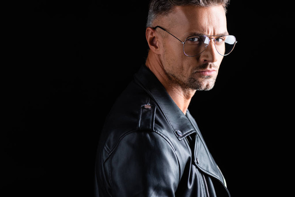 Side view of serious man in sunglasses and leather jacket looking at camera isolated on black  - Valokuva, kuva