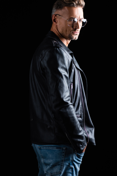 Side view of handsome man in leather jacket and sunglasses looking away isolated on black  - Фото, зображення