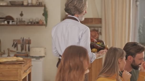 Happy elderly man in apron bringing a baked duck to the table on festive family dinner at home - Imágenes, Vídeo