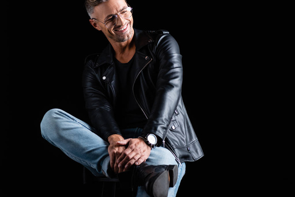 Handsome man in leather jacket and jeans sitting on chair and smiling away isolated on black  - Foto, Imagem
