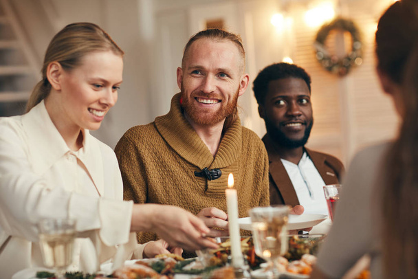 Retrato del hombre barbudo contemporáneo sonriendo felizmente mientras celebra la Navidad con amigos y familiares en la mesa de la cena
 - Foto, Imagen