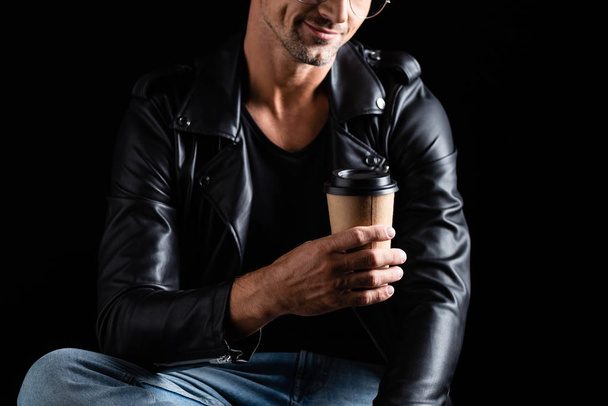 Vista recortada de homem sorridente em jeans e jaqueta de couro segurando copo de papel isolado em preto
  - Foto, Imagem